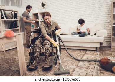 Female Veteran In A Wheelchair Is Cleaning House. Supporting Husband. Camouflage Uniform. Disabled Soldier. Vacuum Cleaner. Working Together. Holiday Leisure. Yellow Gloves. Free Time.