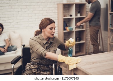 Female Veteran In A Wheelchair Is Cleaning House. Supporting Husband. Camouflage Uniform. Disabled Soldier. Family Concept. Working Together. Holiday Leisure. Yellow Gloves. Free Time.