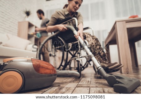 Female Veteran In A Wheelchair Is Cleaning House. Supporting Husband. Camouflage Uniform. Disabled Soldier. Vacuum Cleaner. Working Together. Holiday Leisure. Yellow Gloves. Free Time.