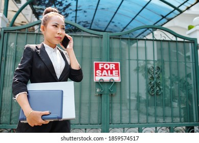 Female Vietnamese Real Estate Agent With Documents In Hands Standing At Gates Of House On Sale And Talking On Phone