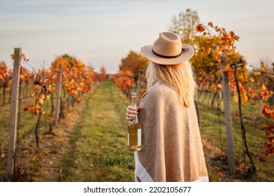 Female vintner enjoying white wine in her vineyard. Woman with poncho and hat holding wine bottle outdoors. Winery at autumn - Powered by Shutterstock