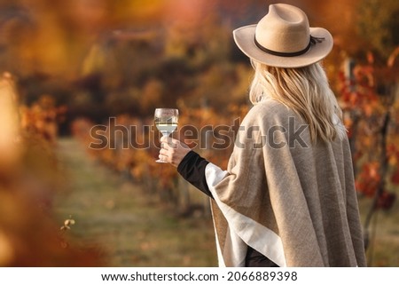 Female vintner enjoying white wine in her vineyard. Woman with poncho and hat holding glass of wine outdoors. Winery at autumn
