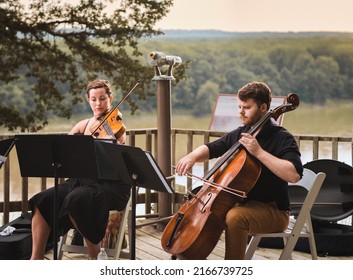 
Female Violin Player And Male Cello Player Playing Duet On Wooden Deck Over Missouri River On Summer Evening