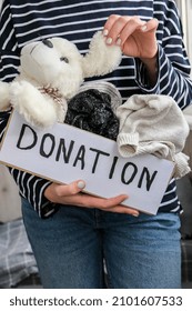 Female Volunteer Holding Donation Box With Old Used Toys And Clothes Indoors. Happy Charity. Unrecognizable Woman Holding Box With Clothes In It. Close-up. Clothing Donation. Winter Clothes 