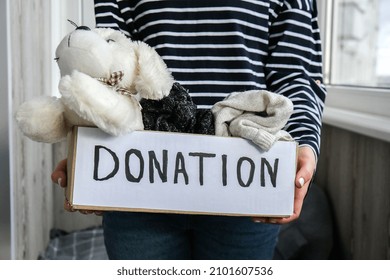 Female Volunteer Holding Donation Box With Old Used Toys And Clothes Indoors. Happy Charity. Unrecognizable Woman Holding Box With Clothes In It. Close-up. Clothing Donation. Winter Clothes 