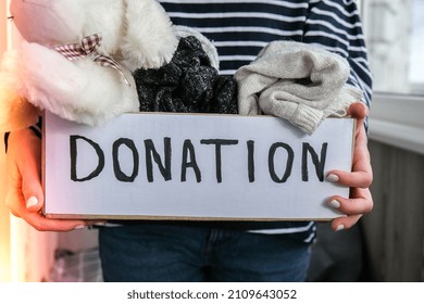 Female Volunteer Holding Donation Box With Old Used Toys And Clothes Indoors. Happy Charity. Unrecognizable Woman Holding Box With Clothes In It. Close-up. Clothing Donation. Winter Clothes 