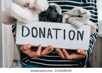 Female Volunteer Holding Donation Box With Old Used Toys And Clothes Indoors. Happy Charity. Unrecognizable Woman Holding Box With Clothes In It. Close-up. Clothing Donation. Winter Clothes 