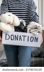 Female Volunteer Holding Donation Box With Old Used Toys And Clothes Indoors. Happy Charity. Unrecognizable Woman Holding Box With Clothes In It. Close-up. Clothing Donation. Winter Clothes 