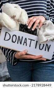 Female Volunteer Holding Donation Box With Old Used Toys And Clothes Indoors. Happy Charity. Unrecognizable Woman Holding Box With Clothes In It. Close-up. Clothing Donation. Winter Clothes 