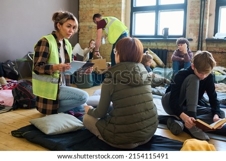 Female volunteer with mobile gadgets communicating to one of refugees while sitting on squats in front of her in shelter for migrants