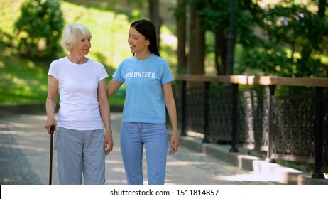 Female Volunteer And Senior Patient With Stick Walking Hospital Garden, Care