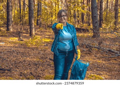 A female volunteer smiles while standing in the forest with a garbage bag in her hand and points her finger at the camera. The woman urges that you too can help save nature from garbage. - Powered by Shutterstock