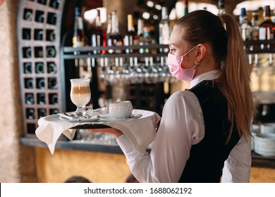 A female Waiter of European appearance in a medical mask serves Latte coffee