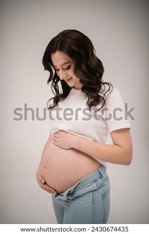 Female waiting for newborn baby on white background. Young pregnant girl touching and holding belly and caring about health indoors. Pregnancy motherhood procreation concept. Belly of a pregnant woman
