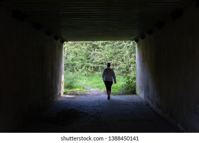 Female walking through an underpass by herself. Dark inside the tunnel, but bright and overgrown on the exit. She is wearing a white jacket and dark trousers. Tunnel is constructed from concrete