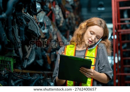 Female warehouse worker checking inventory of used car engine parts while talking on smartphone, industrial inspection and stock management process in automotive recycling storage facility