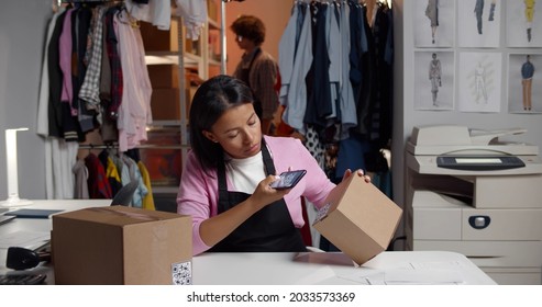 Female warehouse worker holding phone scanning retail package qr-code. Portrait of african woman working in modern online clothes store warehouse