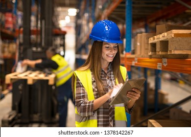 Female Warehouse Worker Holding Tablet Checking Inventory In Distribution Warehouse. Forklift In Background.