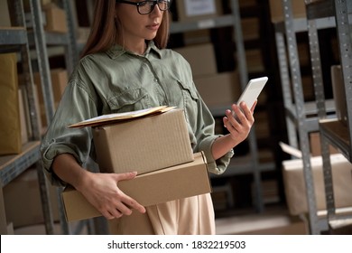 Female Warehouse Worker Manager, Small Stock Business Owner Holding Phone And Retail Package Parcel Boxes Checking Commercial Shipping Delivery Order On Smartphone Using Mobile App Technology.