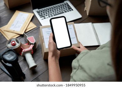 Female warehouse worker, seller, dropshipping small business owner holding phone mock up screen ad scan retail package postal parcel bar code on commercial shipping box on smartphone using mobile app.