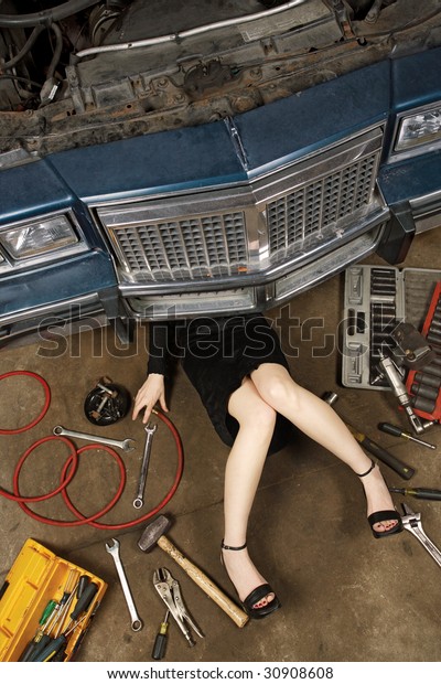 A female wearing\
a black skirt and heels doing repairs under the front of an old car\
from the early 80\'s.