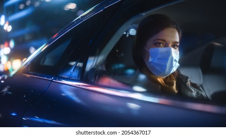 Female Wearing Face Mask is Commuting Home in Backseat of a Taxi at Night. Beautiful Passenger Looking Out of Window while in a Car in Urban City Street with Working Neon Signs. - Powered by Shutterstock