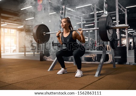 Female weightlifter is getting ready to stand with heavy barbell, squatting with weight, looking away with pleasant expression, enjoying training in gym, portrait, side shot