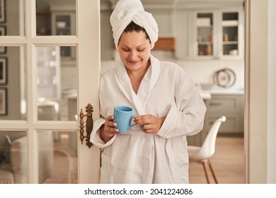 Female in white bathrobe and with towel at her head is standing on the living room