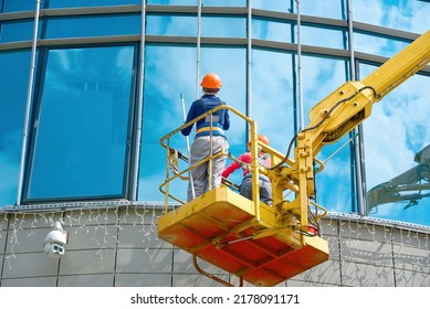 Female Window Cleaner Cleaning Glass Windows On Building At Height In Lifting Platform. Worker Polishing Glass, Window Washing On Office Building In Crane Bucket. Window Cleaners Exterior Glass Facade