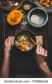 Female Women Hands Holding Pot And Spoon And Cooking Pumpkin Risotto On Dark Rustic Kitchen Table With Ingredients, Top View. Clean Seasonal Food And Eating 