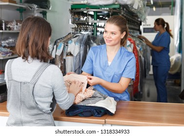 Female Worker Of Dry Cleaner Checking Clothes Of Customer On Reception
