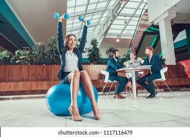 Female Worker Exercising On Fitness Ball In Office.