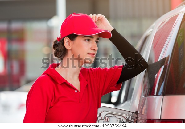 Female\
worker at gas station. Young assistant woman refuelling car at\
petrol station and wear red uniform and red\
hat