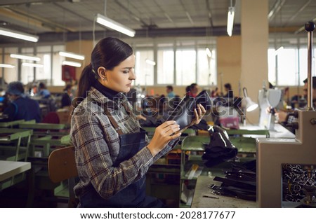 Female worker making new boots standing at a table in shoe factory workshop. Serious young woman in apron checking quality of leather details. Manufacturing industry and footwear production concept