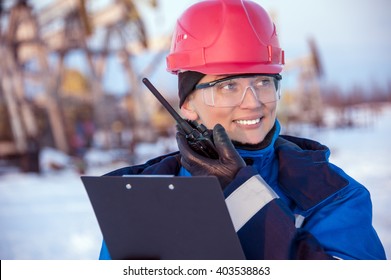 Female worker in the oil field talking on the radio wearing red helmet and blue work clothes. Industrial site background.
