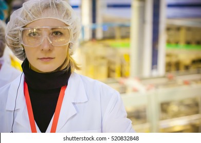 Female Worker On The Production Line Of Beer Factory. Portrait Of A Woman In A White Robe, Standing On The Background Of The Production Line At The Factory Bottling Of Finished Products