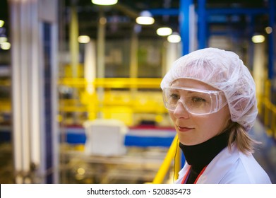 Female Worker On Production Line Of Beer Factory.portrait Woman In White Robe, Standing On Background Of Production Line At Factory Bottling Finished Product, Observing Working Process Through Goggles