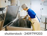 Female worker in uniform and hat using hose while cleaning big factory container for fermenting cheese during workday