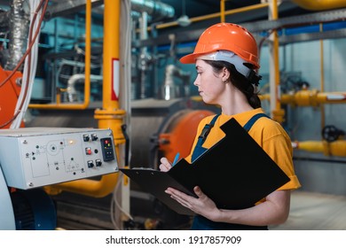 A female worker in uniform and helmet with a folder and a pen in her hand, writing meter readings. In the background-boiler room. Industrial production. - Powered by Shutterstock