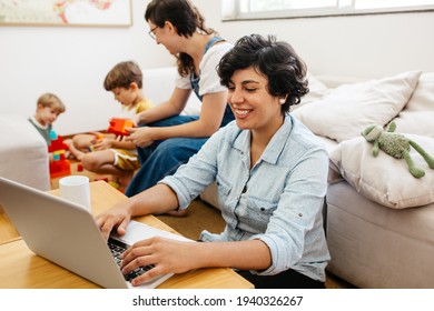 Female Working From Home With Her Family Playing In Background. Lesbian Family Working And Playing With Children.