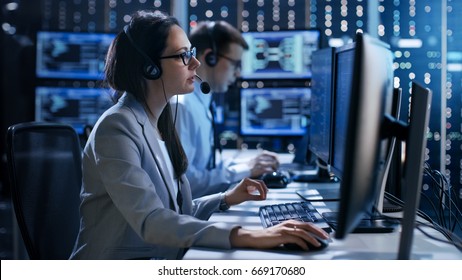 Female working in a Technical Support Team Gives Instructions with the Help of the Headsets. In the Background People Working and Monitors Show Various Information. 