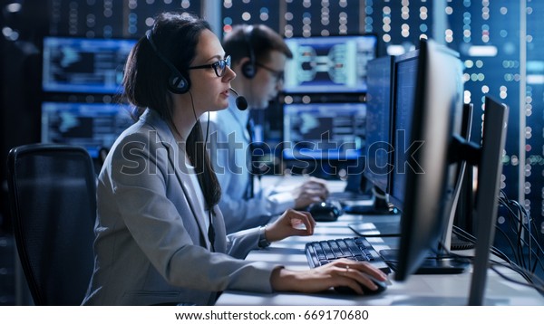 Female working in a Technical\
Support Team Gives Instructions with the Help of the Headsets. In\
the Background People Working and Monitors Show Various\
Information. 