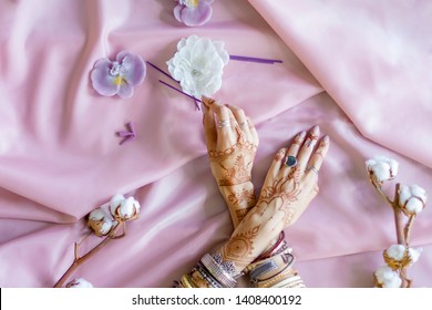 Female wrists painted with traditional Indian oriental mehndi ornaments. Hands dressed in bracelets and rings hold aroma stick. Pink fabric with folds, cotton branches and candles on background.