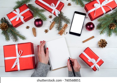 Female Writing Goals To Blank Notebook, Christmas Gifts, Copy Space. Mom Writes To Do List Of Gifts For Christmas And New Year. Top View, Flat Lay. Mobile Phone With White Screen For App Presentation