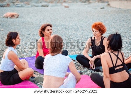 Female yoga class. Group of happy young-adult multi-racial women are sitting on sports mats on wild beach and talking to each other. Concept of female circle of communication and support.