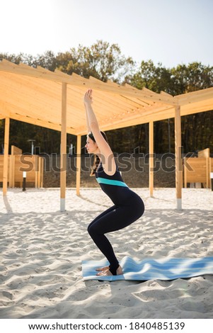 A female yogi performs the Utkatasana asana on a blue Mat on a sandy beach