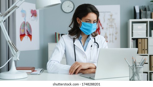 Female Young Doctor Showing To Camera How Wear Medical Mask While Sitting At Desk With Laptop Computer. Medical Vlog Concept.