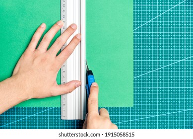 Female young hands cutting a green sheet of paper with a cutter and a metal ruler on a cutting board. Flat lay image with copy space.