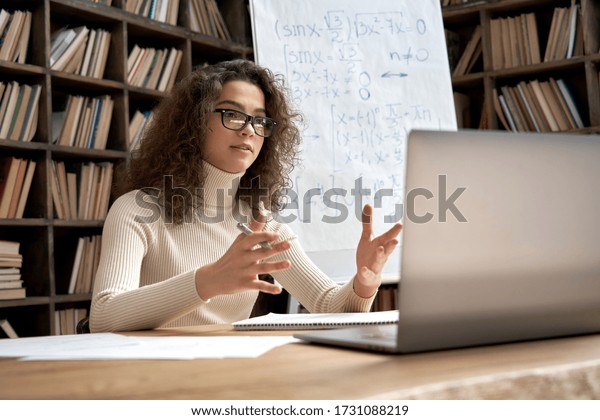 Female young hispanic school math teacher,\
college tutor coach looking at webcam and talking in classroom\
giving virtual teaching remote class online lesson by zoom\
conference call on laptop\
computer.