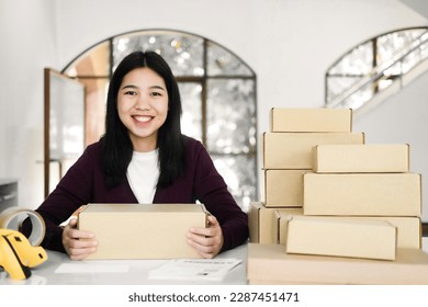 Female young online business, store owner or entrepreneur sitting at desk with cardboard boxes, scanner, and tape holding parcel and looking at camera with happy and smiley face. Online business - Powered by Shutterstock